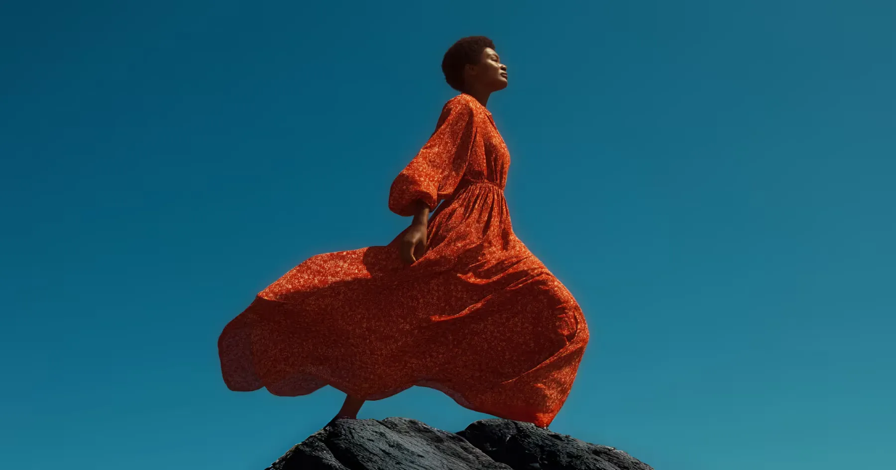 A person in flowing orange clothing posed on a rock against a teal background